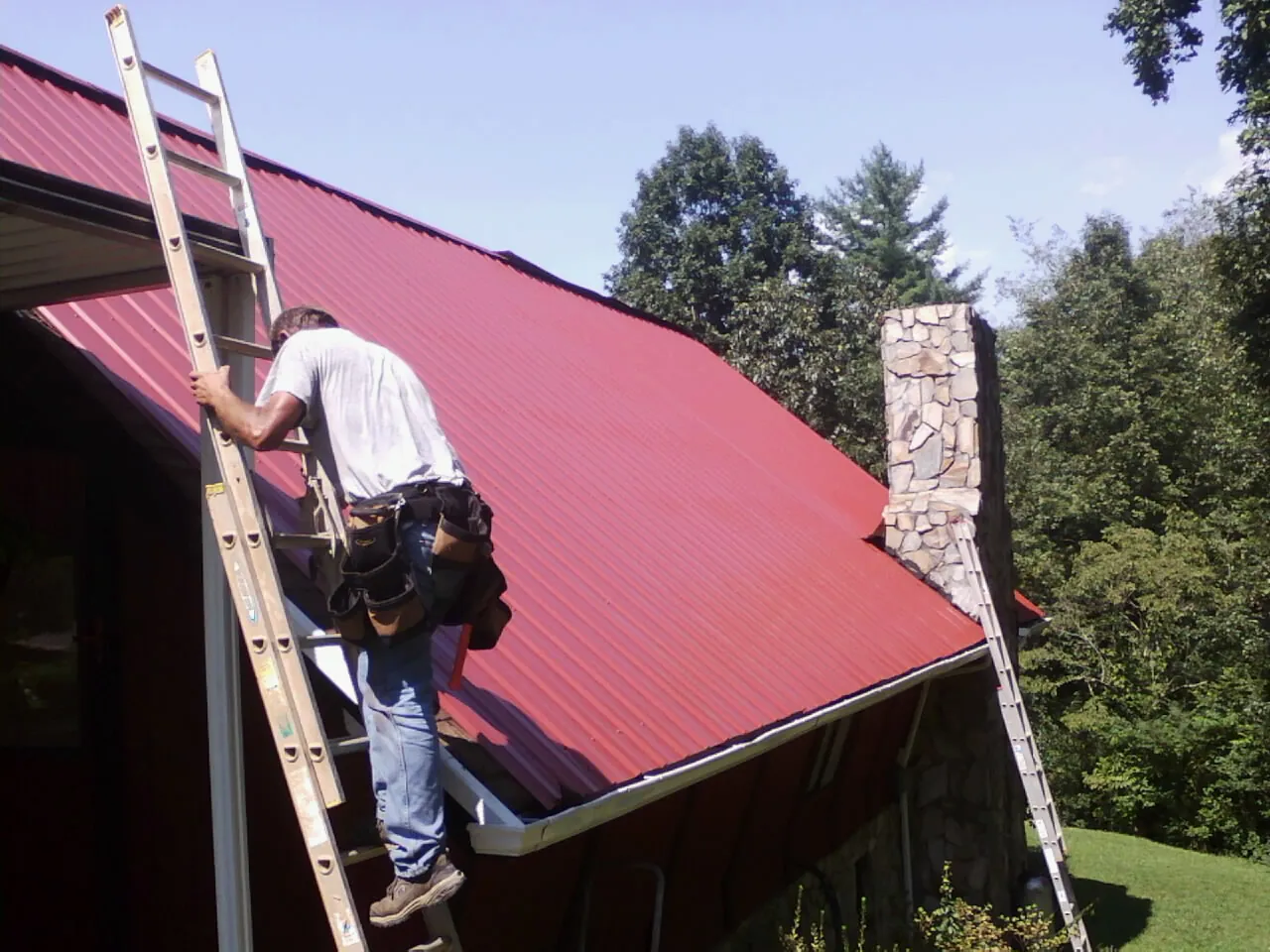 Roofer on a ladder beside a red metal roof and stone chimney — Soffit & Fascia Repair by Roofing Brothers in Stevensville